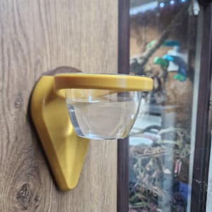 May include: A yellow plastic wall-mounted feeder with a clear plastic bowl. The feeder is designed to hold food or water for small animals.