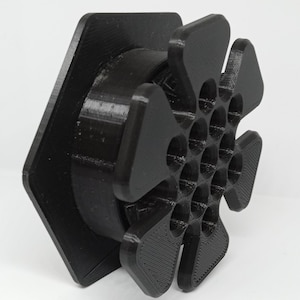 May include: A black plastic hexagonal base with a 3D printed, multi-faceted, star-shaped design. The star has a honeycomb pattern with multiple circular holes.