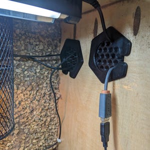 May include: Interior of a reptile enclosure. A black wire mesh lamp enclosure is on the left. Two black hexagonal vents are mounted on the wooden wall. A gray and black USB cable is plugged into one of the vents.