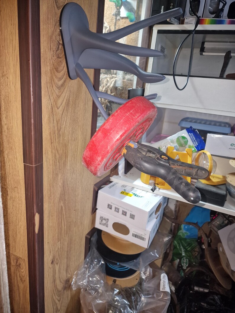 Puede incluir: Un gancho de pared en forma de pulpo de color gris impreso en 3D con cinco brazos. Una placa de peso roja est&aacute; colgando de uno de los brazos. Una abrazadera negra mantiene la placa de peso en su lugar.