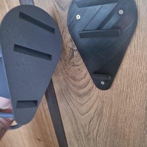 Puede incluir: Dos soportes de pared negros impresos en 3D con una superficie texturizada. Un soporte tiene tres ranuras y el otro tiene dos.