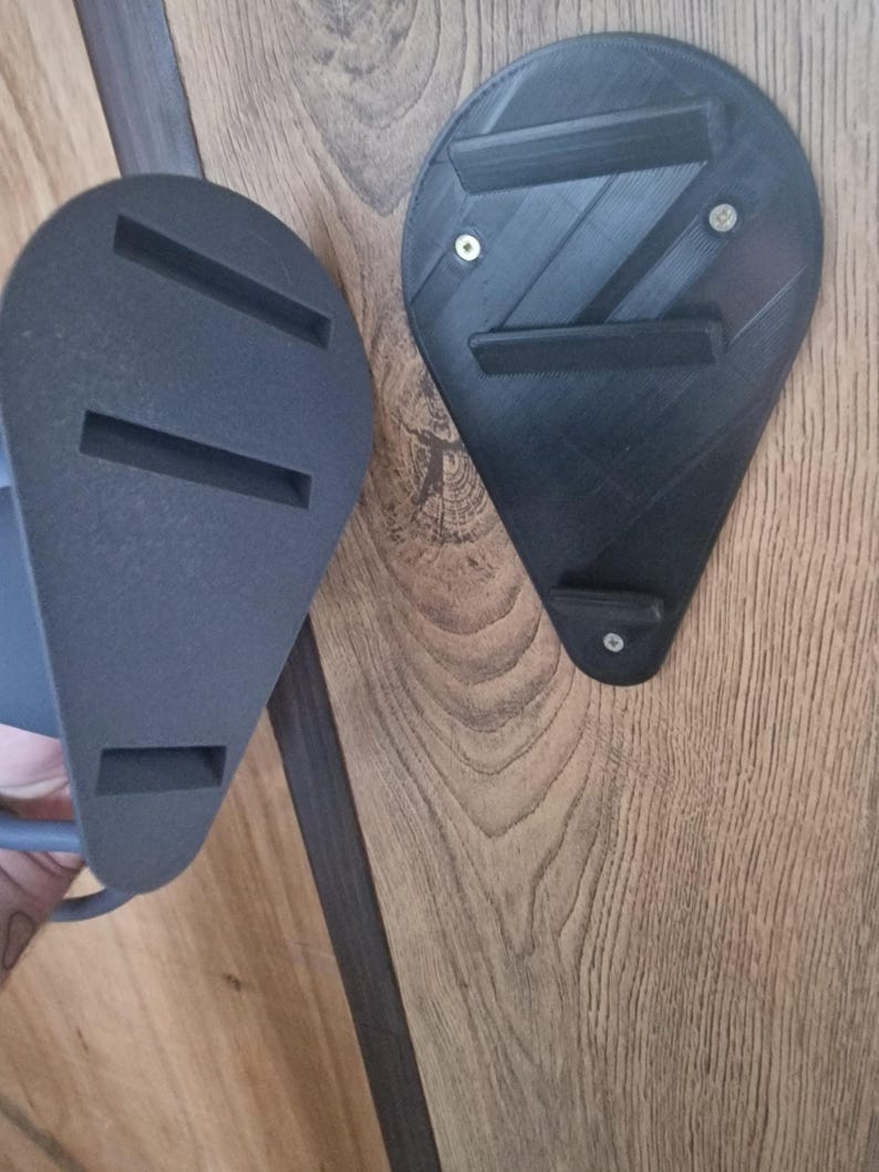 Puede incluir: Dos soportes de pared negros impresos en 3D con una superficie texturizada. Un soporte tiene tres ranuras y el otro tiene dos.