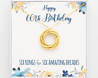 Può includere: Una collana d'oro con un ciondolo a sei anelli in una scatola bianca con un design floreale. La scatola ha il testo "Happy 60th Birthday" e "Six rings for six amazing decades".