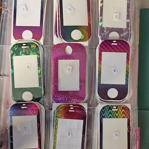 May include: A collection of nine stacks of rectangular adhesive phone skin decals with rounded corners, each featuring a unique holographic or glitter pattern, a central white rectangular cutout, and a circular bottom cutout, organized in a clear plastic binder.