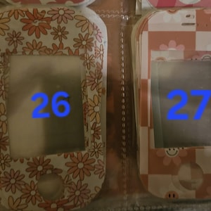 May include: Two decorative cases for a small electronic device. The case on the left has a white background with orange and pink flowers. The case on the right has a pink and white checkered pattern with flowers and a smiling face. The number 26 and 27 are visible.