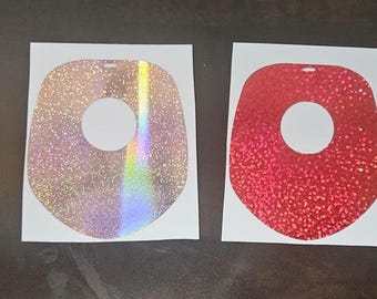 May include: Four glittery, die-cut stickers in a row. The stickers are in the shape of a rounded square with a hole in the center. Colors include gold, iridescent, red, and copper. Each sticker is on a white backing.