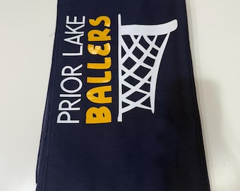 May include: A dark navy blue rectangular fabric banner featuring the white text PRIOR LAKE and the yellow text BALLERS, accompanied by a white graphic illustration of a basketball hoop net positioned to the right of the lettering.