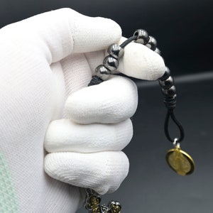 May include: A black beaded bracelet with a gold pendant. The bracelet is made of small, round beads and has a simple design. The pendant is a round disc with a design on it.