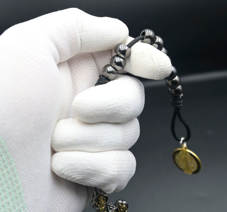 May include: A black beaded bracelet with a gold pendant. The bracelet is made of small, round beads and has a simple design. The pendant is a round disc with a design on it.
