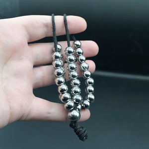 May include: A silver beaded necklace with three strands of beads. The beads are round and smooth, and the necklace is strung on a black cord.