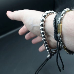 May include: A silver beaded bracelet with a black cord and a black leather bracelet with a silver chain and gold accents.