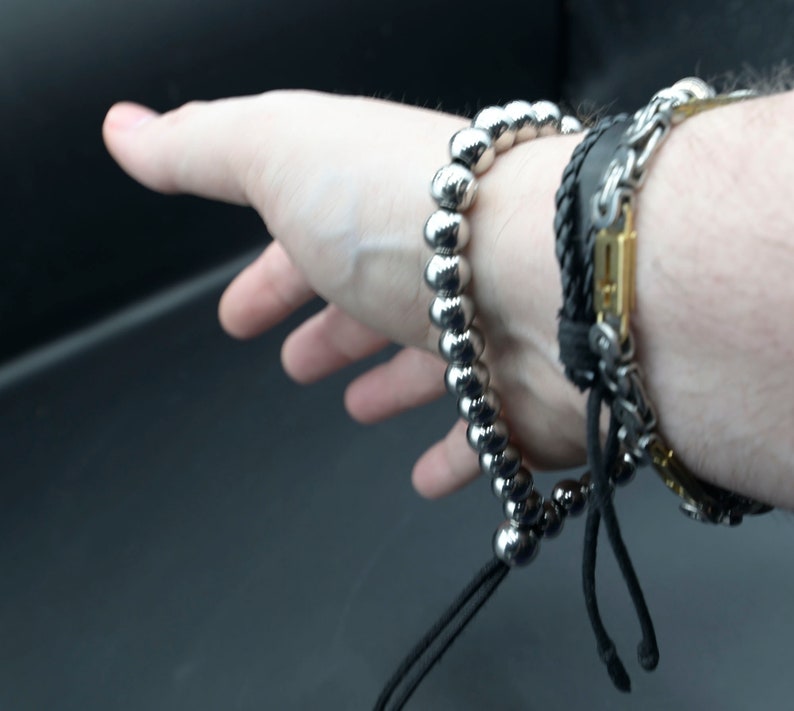 May include: A silver beaded bracelet with a black cord and a black leather bracelet with a silver chain and gold accents.