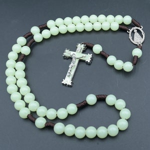 May include: A glow-in-the-dark rosary with brown string and white beads. The rosary features a silver crucifix with a figure of Jesus Christ and a silver pendant with a religious image.