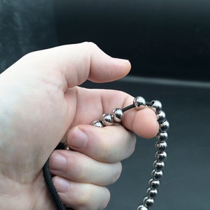 May include: A silver beaded bracelet with a black string threaded through the beads. The bracelet is being held in a hand.