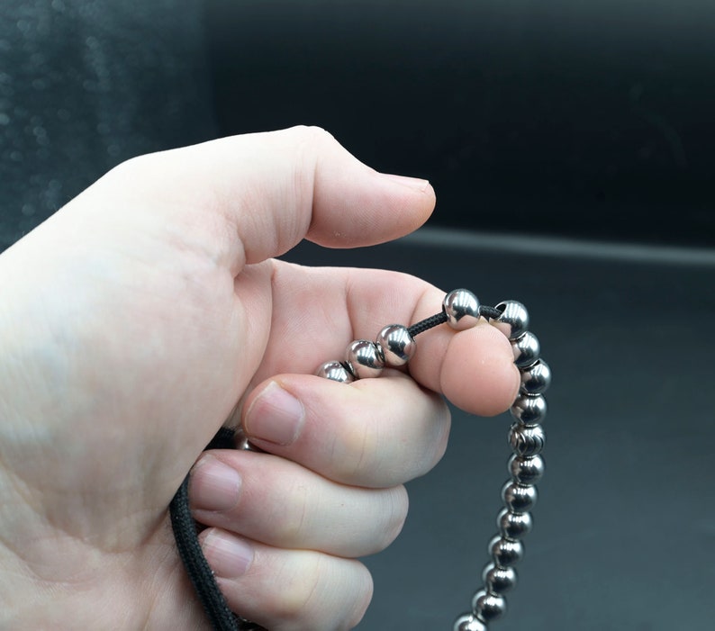 May include: A silver beaded bracelet with a black string threaded through the beads. The bracelet is being held in a hand.
