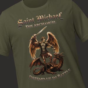 May include: An olive green t-shirt featuring a graphic of Saint Michael the Archangel with large wings, wearing armor, and holding a sword while standing over a dragon. The text reads Saint Michael The Archangel and Defend Us In Battle.