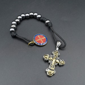May include: A black cord bracelet with silver beads and a silver cross pendant. The cross has a gold-colored center with a figure of Jesus Christ. The bracelet also has a small round pendant with a red and blue design.