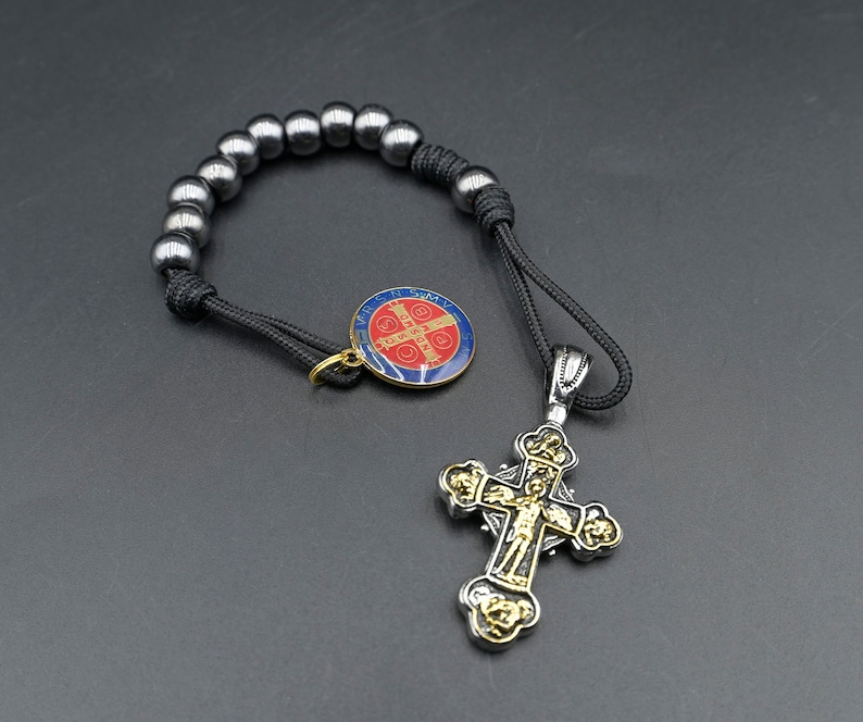 May include: A black cord bracelet with silver beads and a silver cross pendant. The cross has a gold-colored center with a figure of Jesus Christ. The bracelet also has a small round pendant with a red and blue design.