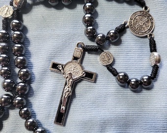Puede incluir: Un rosario con cuentas esféricas de color gris oscuro y un cordón negro. El rosario presenta un crucifijo plateado con una representación detallada de Jesucristo y varias medallas plateadas. La cruz es negra con detalles plateados.