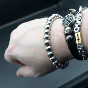 May include: A silver beaded bracelet with a black leather bracelet with a silver cross charm and a silver charm with a black design.