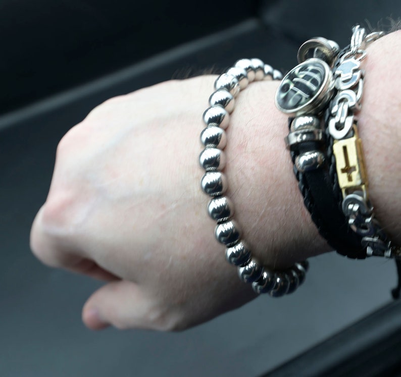 May include: A silver beaded bracelet with a black leather bracelet with a silver cross charm and a silver charm with a black design.