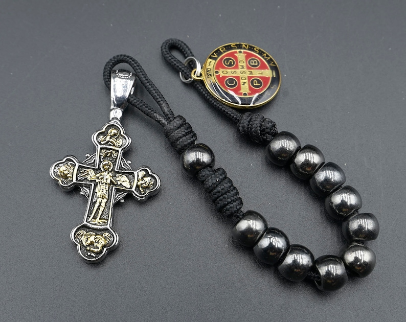 May include: A black cord bracelet with black beads and a silver cross pendant. The cross has gold accents and a small figure of Jesus on it. The bracelet also has a small round metal charm with the words "VR.S.N.S.M.V." and a red and white cross.