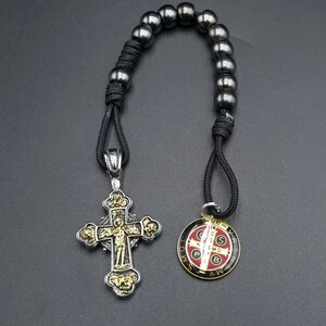 May include: A black cord rosary with a silver and gold cross pendant and a round silver and red medal pendant. The medal has the inscription "S.S.M.D. P.B. A.W.S.V.R.S."