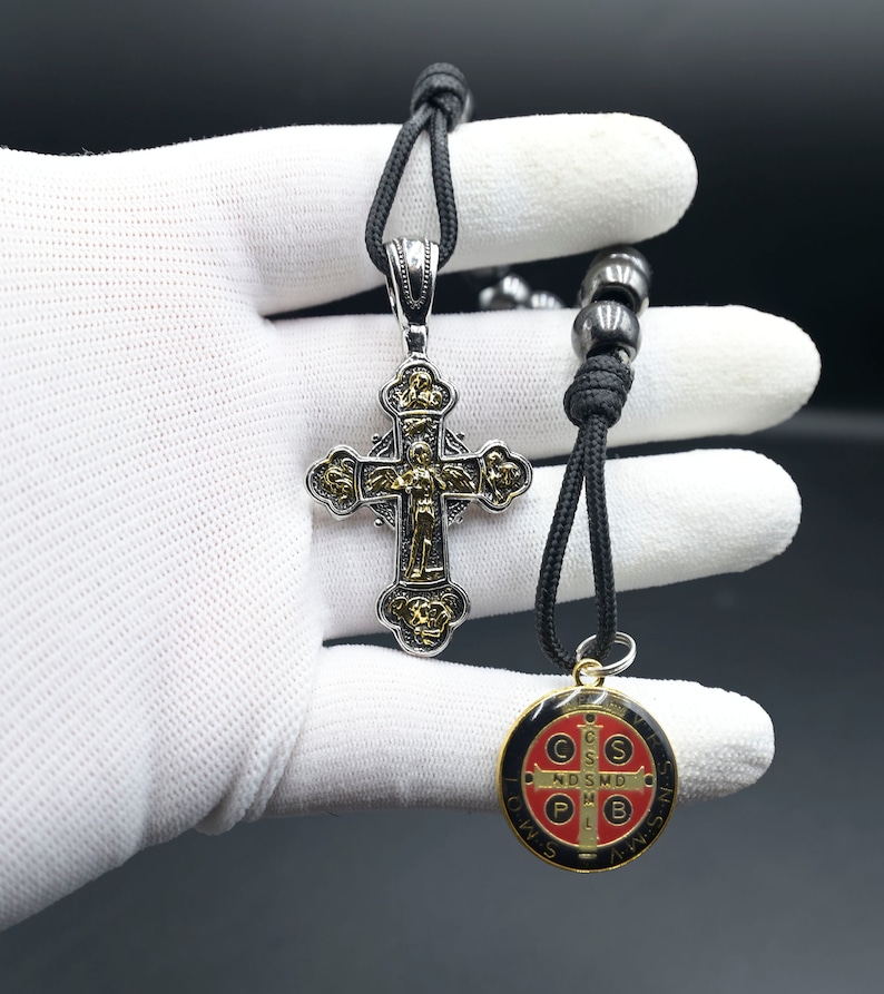 May include: A black cord necklace with a silver and gold cross pendant and a round, red, black, and gold medal pendant. The medal has the words "S.P.Q.R." and "O.W.S.S.N.S." around the edge.
