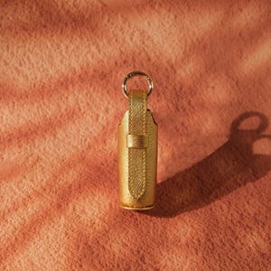 May include: A metallic gold cylindrical case with a vertical strap and a circular metal ring attached to the top. The exterior features a textured, shimmering finish, and the design includes a small loop to secure the strap against the main body.