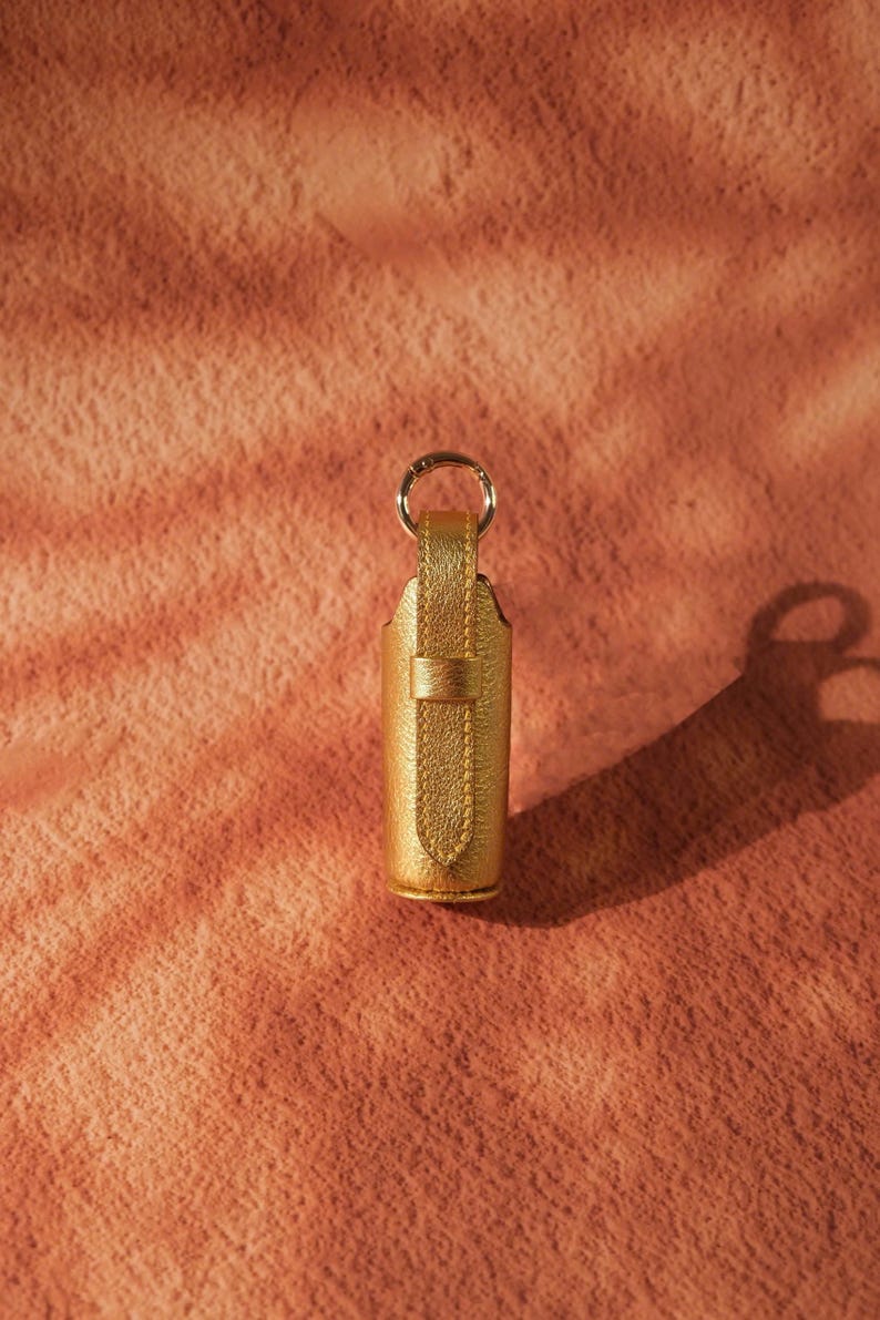 May include: A metallic gold cylindrical case with a vertical strap and a circular metal ring attached to the top. The exterior features a textured, shimmering finish, and the design includes a small loop to secure the strap against the main body.