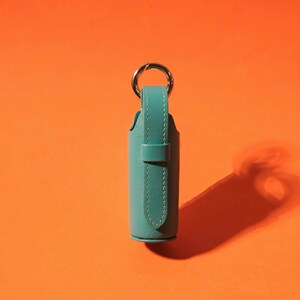 May include: A cylindrical teal leather case featuring a vertical strap with visible stitching and a silver-toned metal ring attached to the top. The case has a smooth texture and a secure loop closure, designed to hold small items securely in place.