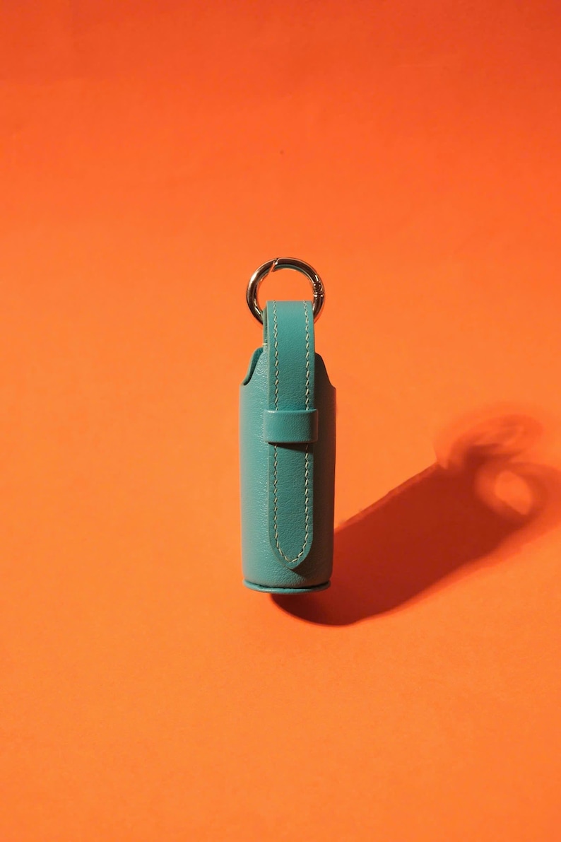 May include: A cylindrical teal leather case featuring a vertical strap with visible stitching and a silver-toned metal ring attached to the top. The case has a smooth texture and a secure loop closure, designed to hold small items securely in place.