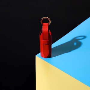 May include: A cylindrical red leather case with a vertical strap and a gold-toned metal ring attached to the top. The case features visible stitching along the edges of the leather strap and body, maintaining a structured and upright vertical orientation.