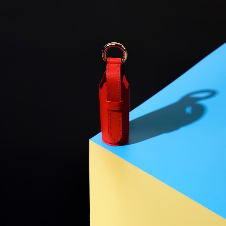 May include: A cylindrical red leather case with a vertical strap and a gold-toned metal ring attached to the top. The case features visible stitching along the edges of the leather strap and body, maintaining a structured and upright vertical orientation.