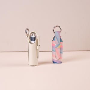 May include: Two cylindrical holders with silver-toned metal key rings. One holder is solid off-white, while the other features a multi-colored diagonal striped pattern in pastel shades of pink, blue, yellow, and purple. Both have a vertical strap.