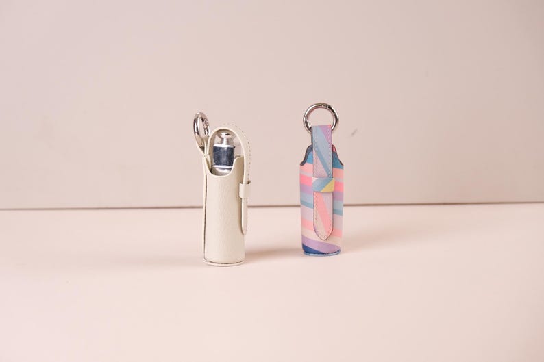 May include: Two cylindrical holders with silver-toned metal key rings. One holder is solid off-white, while the other features a multi-colored diagonal striped pattern in pastel shades of pink, blue, yellow, and purple. Both have a vertical strap.