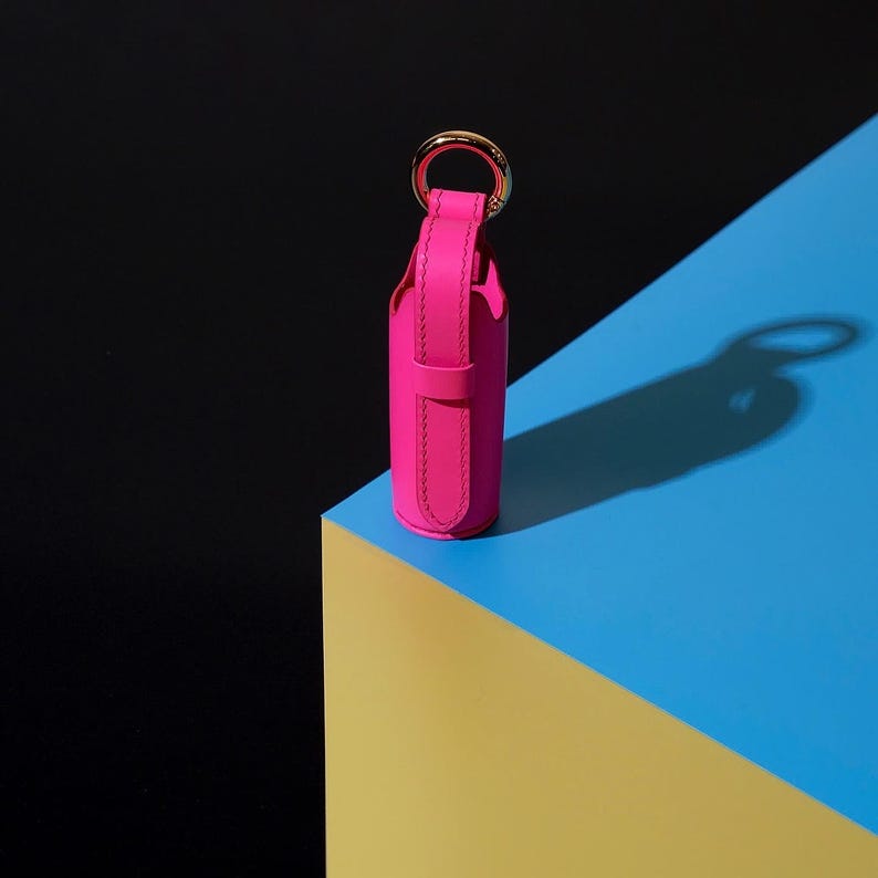 May include: A bright hot pink leather cylindrical case featuring a vertical strap closure and a polished gold-tone metal ring attached to the top. The accessory has visible stitching along the edges and a secure loop to hold the strap in place.