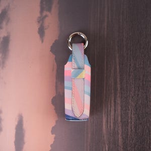 May include: A vertical rectangular leather holder featuring a diagonal striped pattern in pastel shades of pink, blue, yellow, and lavender. The holder includes a silver-toned metal circular ring attached to the top for hanging or attachment purposes.
