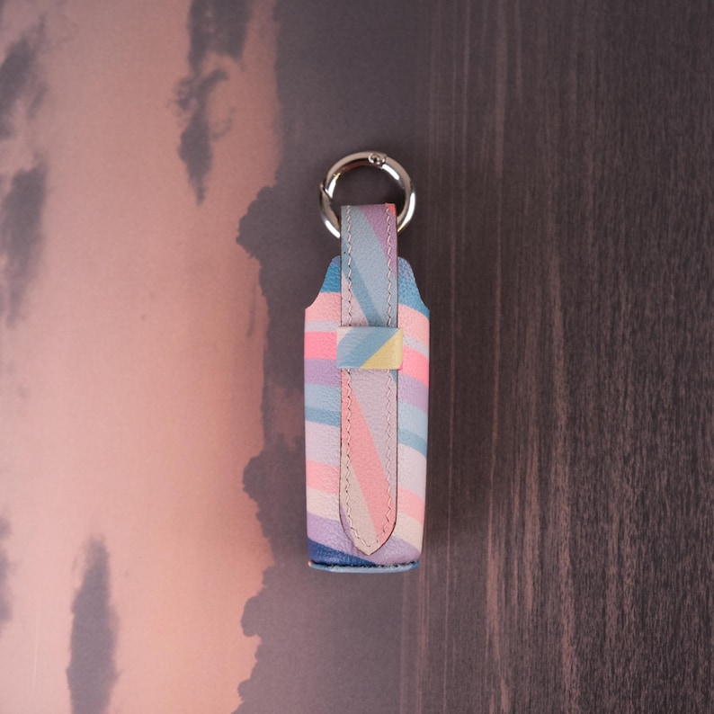May include: A vertical rectangular leather holder featuring a diagonal striped pattern in pastel shades of pink, blue, yellow, and lavender. The holder includes a silver-toned metal circular ring attached to the top for hanging or attachment purposes.