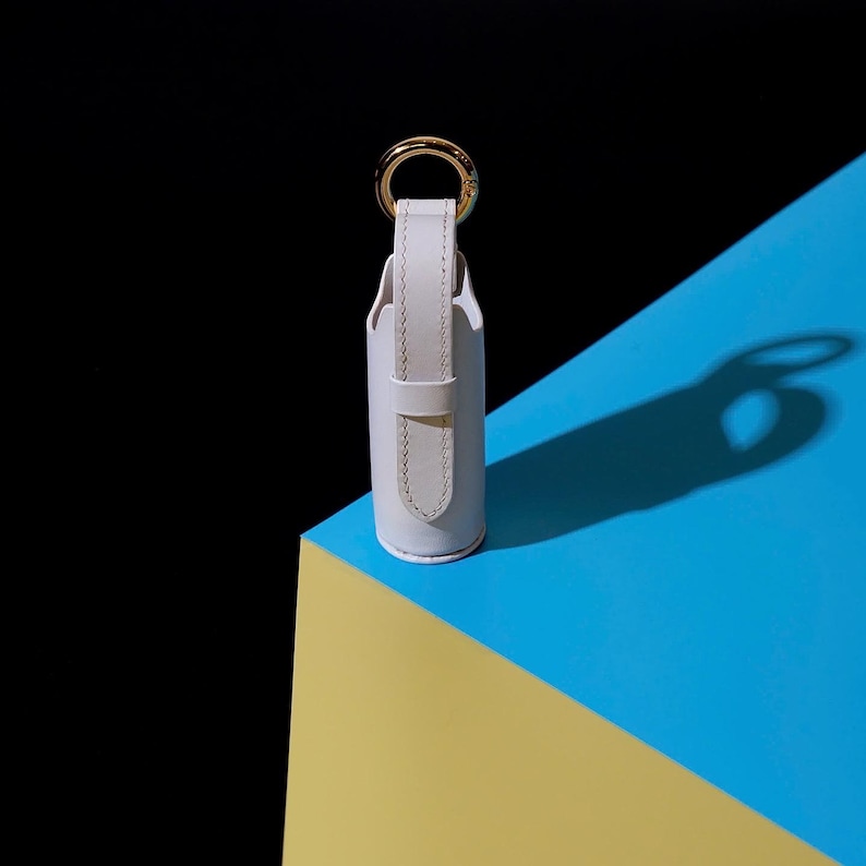May include: A cylindrical white leather holder featuring a vertical strap and a gold-toned metal ring at the top. The holder has a smooth, matte finish with visible stitching along the edges of the strap, providing a structured and clean appearance.