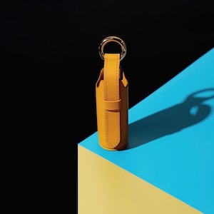 May include: A cylindrical mustard yellow leather case featuring a vertical strap and a gold-toned metal ring at the top. The case has visible stitching along the edges of the strap and a smooth, matte finish, standing upright with a circular base.