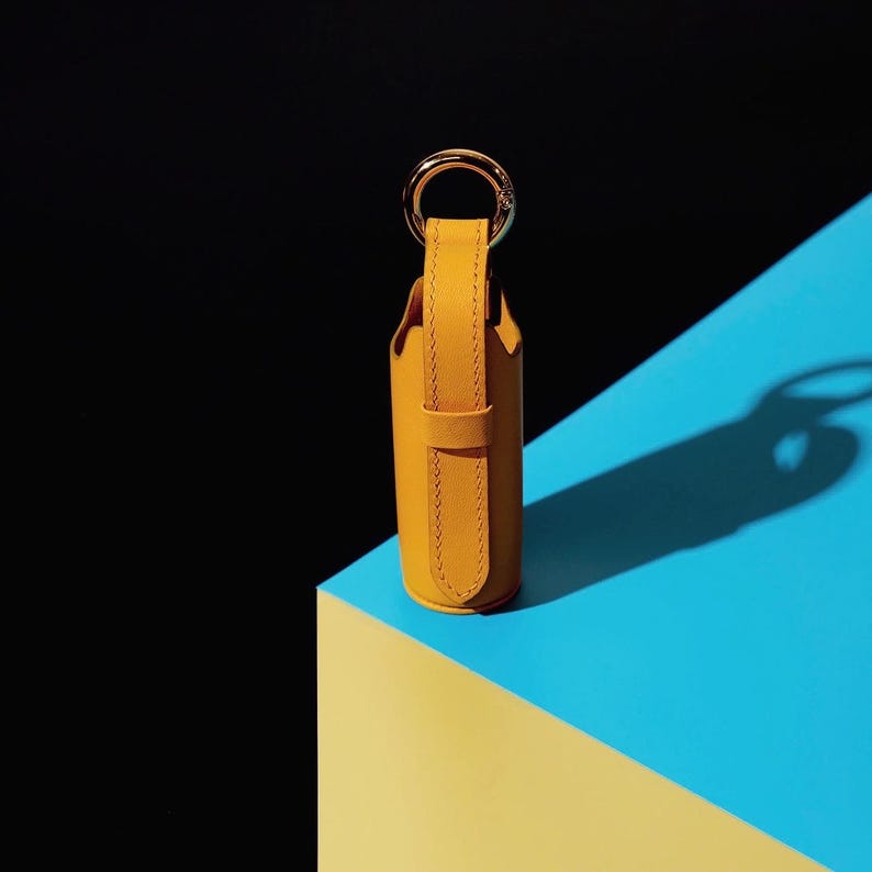 May include: A cylindrical mustard yellow leather case featuring a vertical strap and a gold-toned metal ring at the top. The case has visible stitching along the edges of the strap and a smooth, matte finish, standing upright with a circular base.