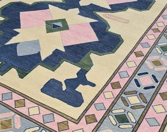 May include: A large, rectangular rug with a cream-colored background and a geometric pattern. The design features navy blue, pink, green, and light blue shapes. The border has a repeating pattern of diamonds and geometric shapes.