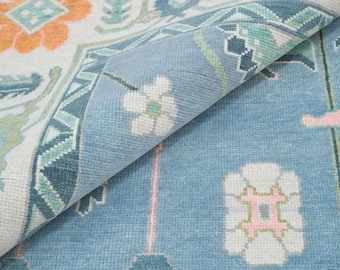 Könnte beinhalten: Nahaufnahme eines aufgerollten Teppichs mit floralem Muster. Der Teppich hat einen hellblauen Hintergrund mit weißen, orangefarbenen und türkisfarbenen floralen und geometrischen Designs. Der gerollte Abschnitt zeigt die Textur des Teppichs.