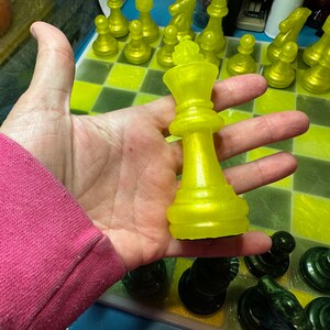 Pu&ograve; includere: Un pezzo del re degli scacchi giallo con una finitura lucida &egrave; tenuto in una mano sopra una scacchiera gialla e verde. Il pezzo del re &egrave; su una casella nera.