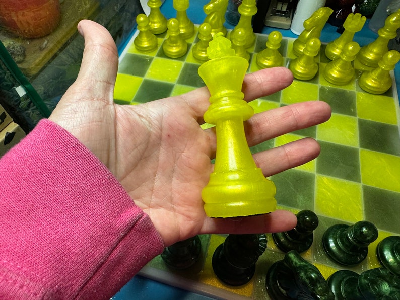 Pu&ograve; includere: Un pezzo del re degli scacchi giallo con una finitura lucida &egrave; tenuto in una mano sopra una scacchiera gialla e verde. Il pezzo del re &egrave; su una casella nera.