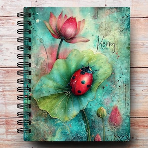 May include: Spiral-bound notebook featuring a watercolor illustration of a red ladybug with black spots on a green lily pad, a pink lotus flower, and the name "Kerry" in cursive. The background is a mix of teal, pink, and green.