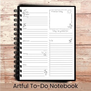May include: A black spiral-bound notebook with a white interior featuring a daily planner layout. The notebook includes sections for the date, morning, noon, afternoon, evening, and notes, along with a "To Do" list and space for gratitude. The text "Artful To-Do Notebook" is at the bottom.