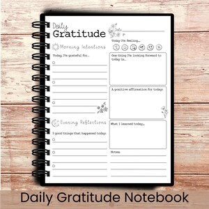 May include: A black spiral-bound notebook with the words "Daily Gratitude" on the top. The notebook has sections for morning intentions, evening reflections, and notes. The bottom of the notebook reads "Daily Gratitude Notebook".