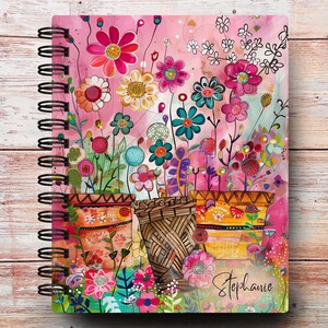 May include: A spiral-bound notebook featuring a vibrant illustration of colorful flowers in three distinct pots against a pink background. The artwork includes various floral shapes and patterns in shades of pink, yellow, teal, and orange. The name Stephanie is written in cursive script in the bottom right corner.